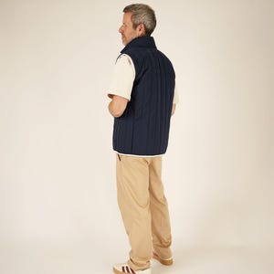 May include: A navy blue quilted vest with white trim is worn over a cream-colored t-shirt. The person is wearing khaki-colored pants and white sneakers with burgundy stripes and a brown sole. The outfit is casual.