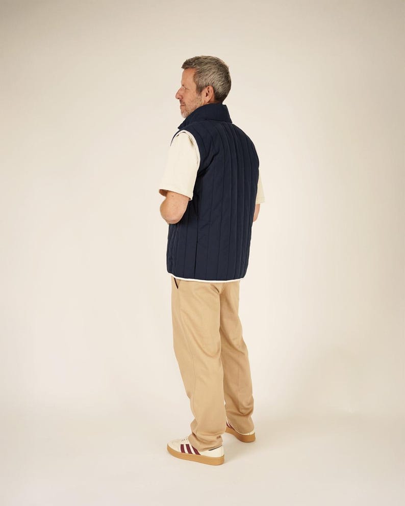 May include: A navy blue quilted vest with white trim is worn over a cream-colored t-shirt. The person is wearing khaki-colored pants and white sneakers with burgundy stripes and a brown sole. The outfit is casual.