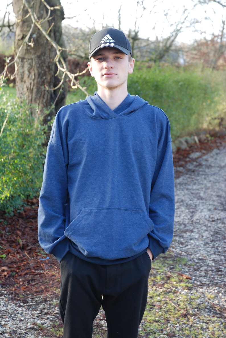 May include: A person wearing a navy blue hoodie with a front pocket and black pants. The person is also wearing a black baseball cap with a white logo. The background includes trees and a path.