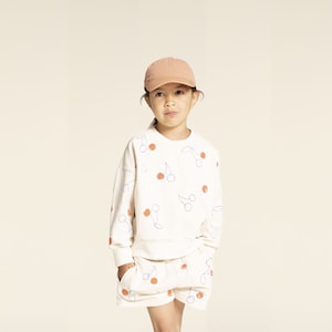 Puede incluir: Una persona joven lleva una sudadera blanca con estampado de cerezas y pantalones cortos blancos a juego con estampado de cerezas. Lleva una gorra de béisbol rosa claro.