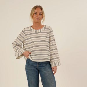 Op de afbeelding: Een crèmekleurige top met lange mouwen en horizontale strepen in zwart, grijs en beige. De top heeft een vierkante halslijn en uitlopende mouwen. De persoon draagt een blauwe jeans.