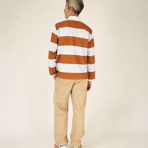 May include: A long-sleeved rugby shirt with horizontal stripes in brown, white, and light blue. The shirt has a white collar and cuffs. The person is wearing khaki-colored pants and sneakers with red and blue accents.