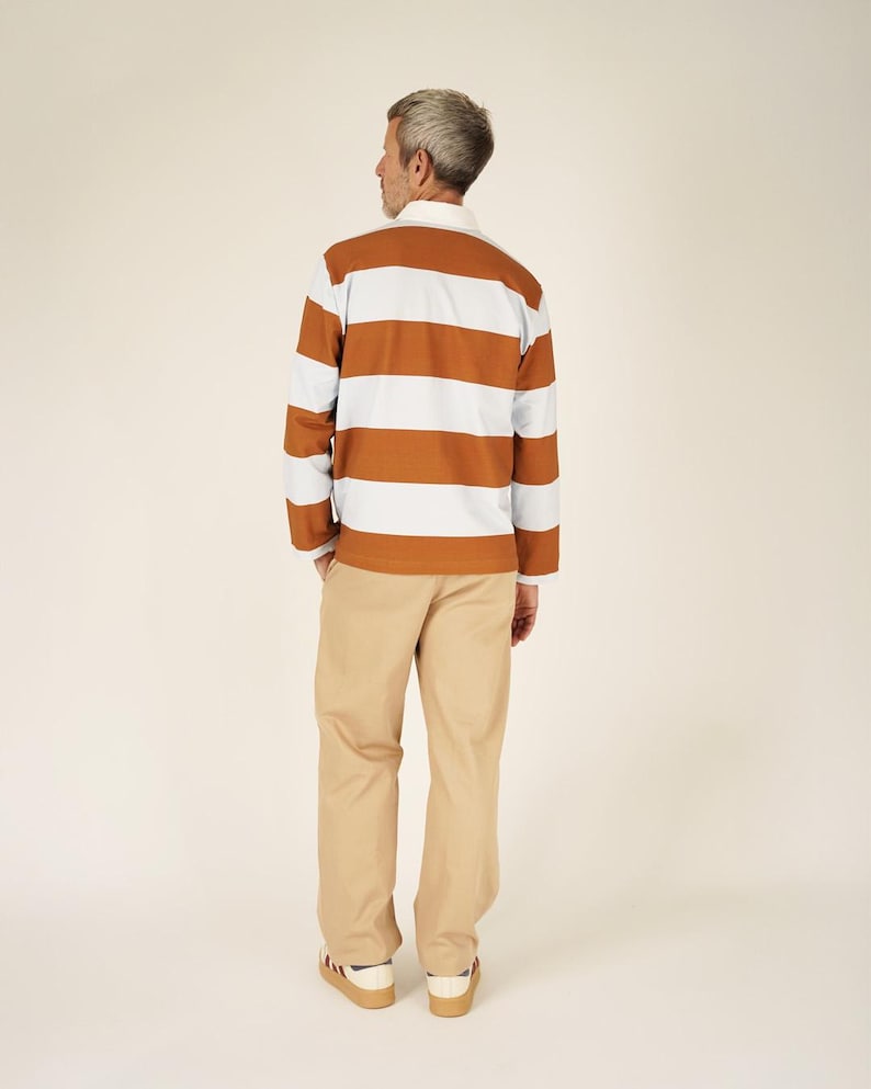 May include: A long-sleeved rugby shirt with horizontal stripes in brown, white, and light blue. The shirt has a white collar and cuffs. The person is wearing khaki-colored pants and sneakers with red and blue accents.