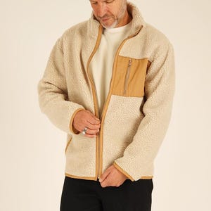 May include: A beige fleece jacket with a tan chest pocket and trim. The jacket has a full zipper closure and a high collar. The person in the image is wearing a yellow beanie and black pants.