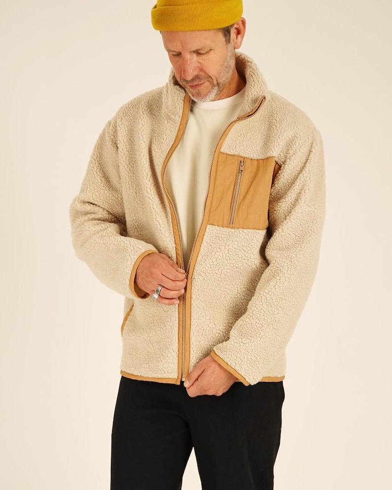 May include: A beige fleece jacket with a tan chest pocket and trim. The jacket has a full zipper closure and a high collar. The person in the image is wearing a yellow beanie and black pants.
