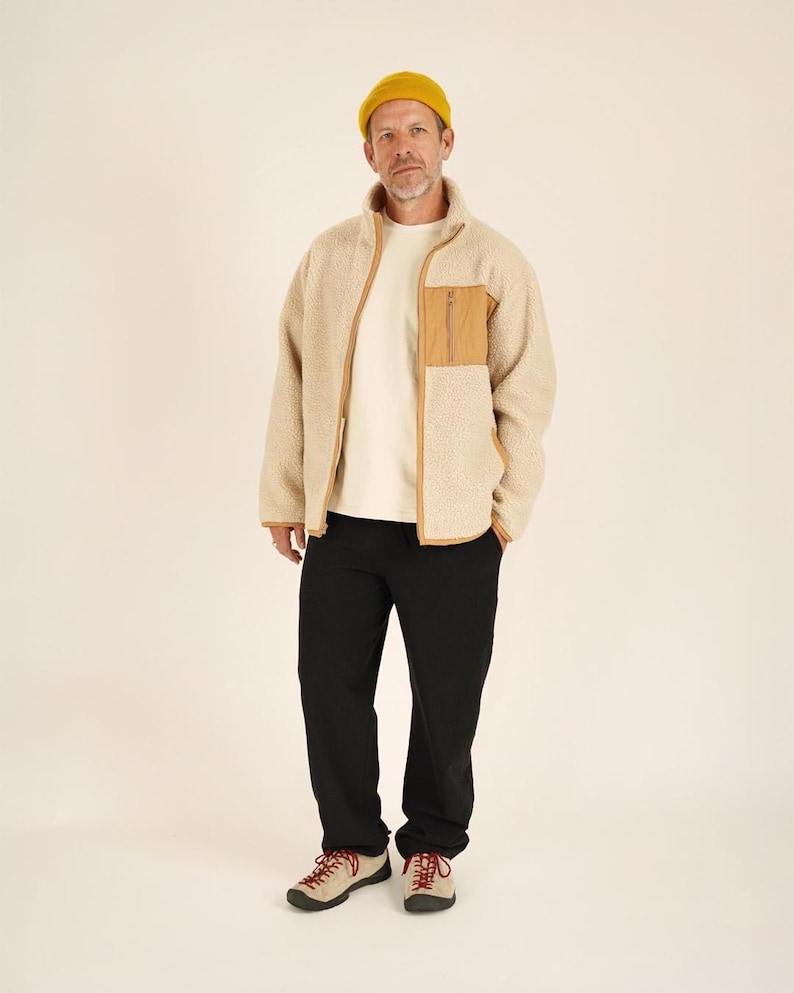 May include: A person wearing a beige fleece jacket with a brown pocket and trim, a white t-shirt, black pants, a yellow beanie, and beige and red sneakers. The jacket has a high collar and a zipper closure.