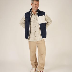 May include: A man wearing a navy blue and white quilted vest over a plaid button-down shirt and khaki pants. He is also wearing white sneakers with burgundy stripes.