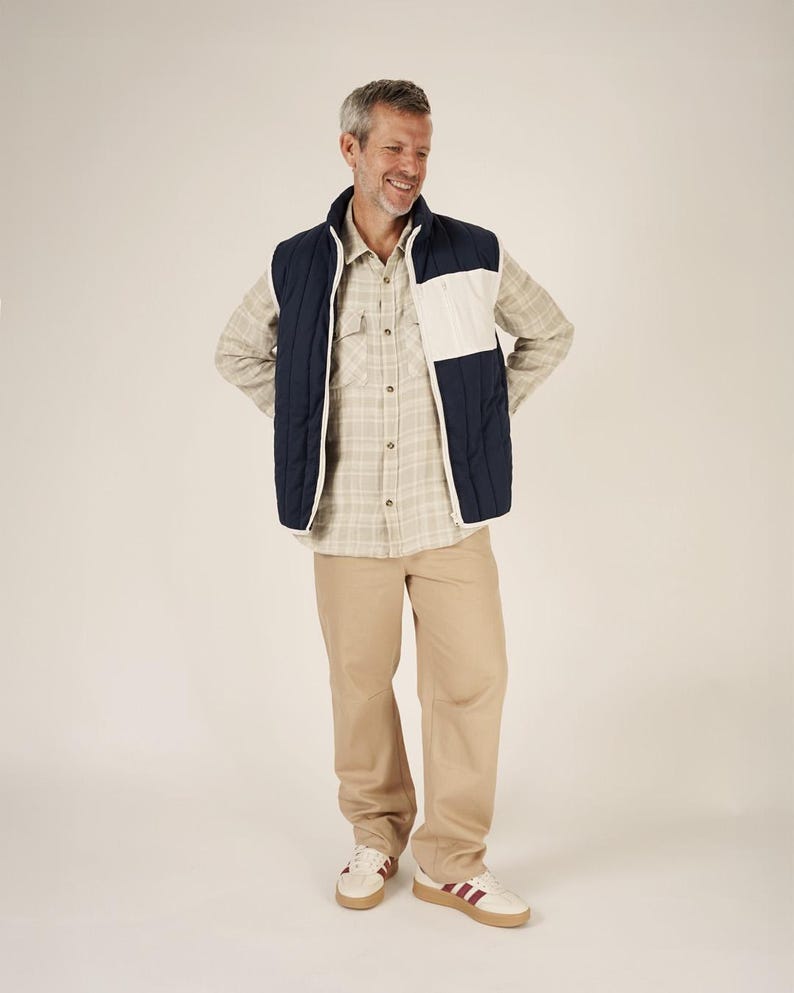 May include: A man wearing a navy blue and white quilted vest over a plaid button-down shirt and khaki pants. He is also wearing white sneakers with burgundy stripes.