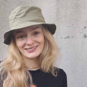 May include: A woman with long blonde hair is wearing a green bucket hat and a black shirt. She is smiling at the camera.