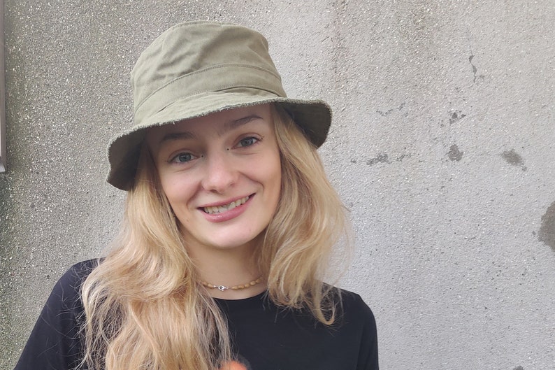 May include: A woman with long blonde hair is wearing a green bucket hat and a black shirt. She is smiling at the camera.