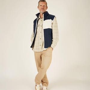 May include: A person wearing a navy blue and white quilted vest over a plaid button-down shirt, paired with khaki pants and cream-colored sneakers with red accents. The vest has a high collar.