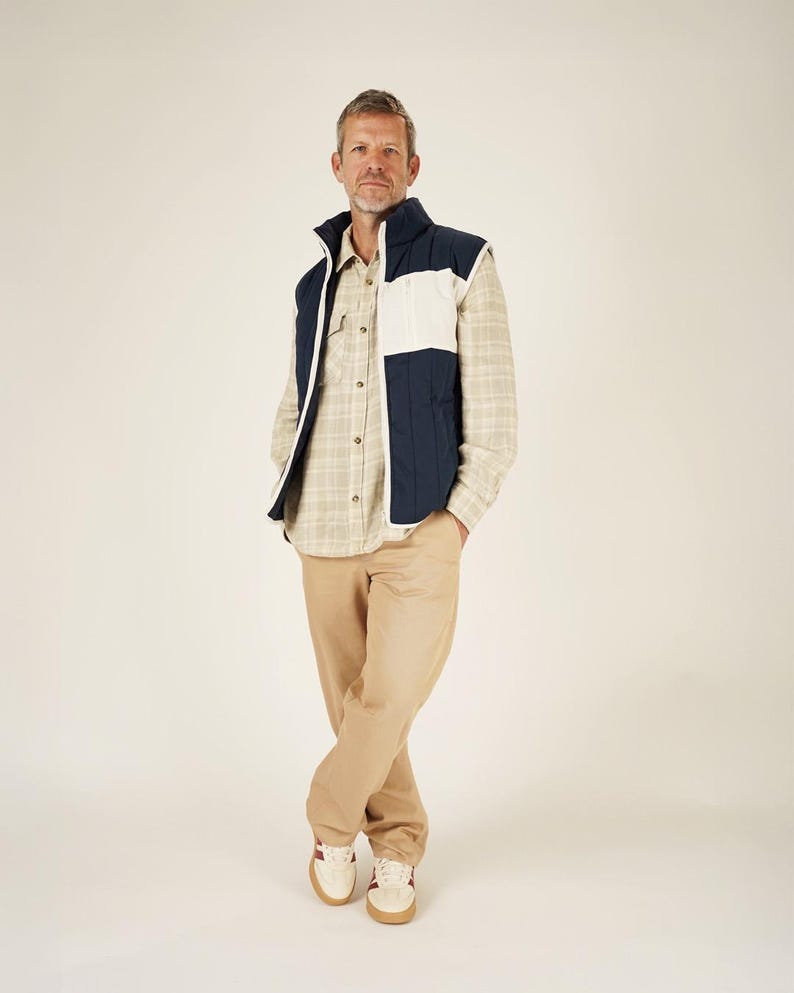 May include: A person wearing a navy blue and white quilted vest over a plaid button-down shirt, paired with khaki pants and cream-colored sneakers with red accents. The vest has a high collar.