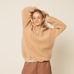 Puede incluir: Una sudadera con capucha de color marrón claro y cierre de cremallera con cuello alto. La sudadera está hecha de un tejido suave y afelpado.