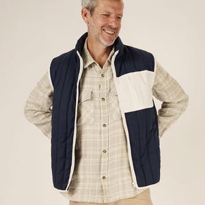 May include: A navy blue and white quilted vest with a full zipper closure. The vest has a white panel on the right side and a high collar. The person in the image is wearing a plaid shirt and khaki pants.