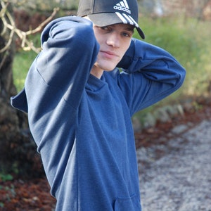 May include: A navy blue hoodie with a front pocket and a black baseball cap with the Adidas logo. The cap has white stripes and a curved brim. The person is wearing the hoodie and cap outdoors.