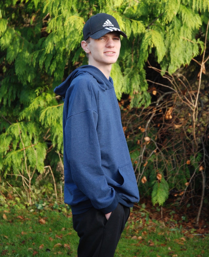 May include: A navy blue hoodie with a front pocket and a black baseball cap with the Adidas logo. The person is wearing black pants and standing in front of green foliage.