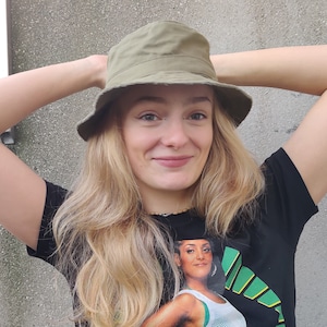 May include: A woman with long blonde hair wearing a green bucket hat and a black t-shirt with a graphic print of a woman in a green and white sports uniform.