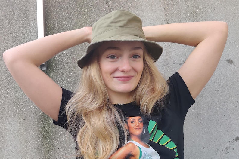 May include: A woman with long blonde hair wearing a green bucket hat and a black t-shirt with a graphic print of a woman in a green and white sports uniform.