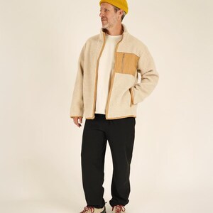 May include: A person wearing a beige fleece jacket with a brown pocket and trim, a white t-shirt, black pants, a yellow beanie, and tan and red hiking shoes. The jacket has a full zipper.