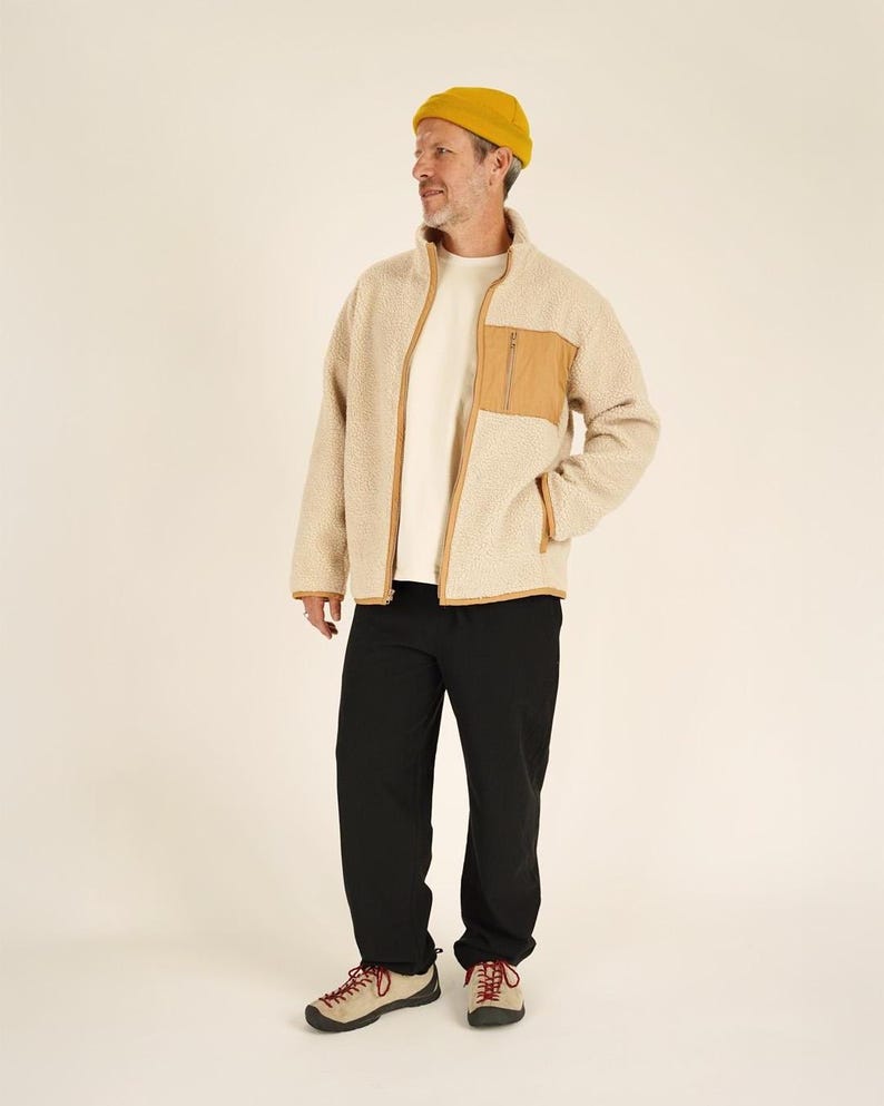 May include: A person wearing a beige fleece jacket with a brown pocket and trim, a white t-shirt, black pants, a yellow beanie, and tan and red hiking shoes. The jacket has a full zipper.