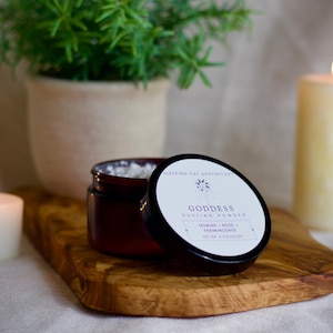May include: A dark amber jar of Goddess dusting powder with a black lid featuring a white label that reads Sleeping Cat Apothecary, Goddess, Jasmine, Rose, Frankincense, and Net Wt 4 ounces, resting on a wooden board with a visible grain.