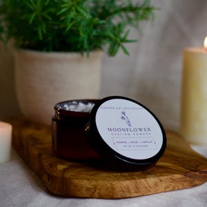 May include: A brown glass jar of Moonflower Dusting Powder with a black lid. The white label features a floral illustration and the text: Sleeping Cat Apothecary, Moonflower Dusting Powder, Jasmine, Rose, Vanilla, Net Wt. 4 Oz (113g).