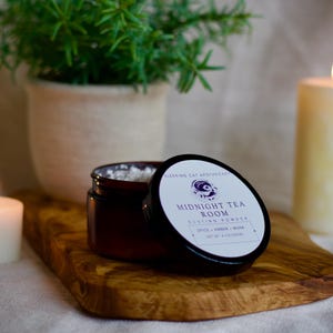 May include: A small amber glass jar containing white dusting powder with a black lid. The label reads Sleeping Cat Apothecary, Midnight Tea Room, Dusting Powder, Spice + Amber + Musk, Net Wt 4 oz (113g). The jar sits on a wooden board.