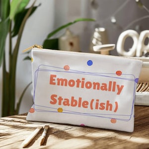May include: A white canvas zipper pouch with the text "Emotionally Stable(ish)" in orange. The design includes a blue outline and colorful polka dots. The pouch has a gold zipper and sits on a wooden surface.
