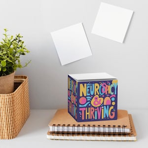 May include: A cube-shaped notepad with the words "Neuro spicy & Thriving" in colorful, playful lettering. The notepad sits on a stack of brown notebooks, with a small potted plant and two blank white sticky notes in the background.