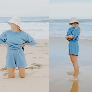 May include: A woman wearing a light blue, oversized, short-sleeved top and matching shorts. She is wearing a white bucket hat and is standing on a sandy beach.
