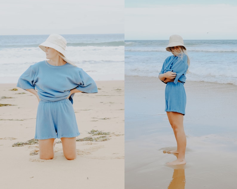 May include: A woman wearing a light blue, oversized, short-sleeved top and matching shorts. She is wearing a white bucket hat and is standing on a sandy beach.