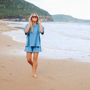 May include: A woman wearing a light blue, short-sleeved,  cotton  set with matching shorts. She is walking on a sandy beach with the ocean in the background. She is wearing sunglasses.