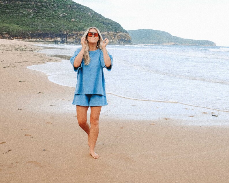May include: A woman wearing a light blue, short-sleeved,  cotton  set with matching shorts. She is walking on a sandy beach with the ocean in the background. She is wearing sunglasses.