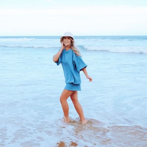 May include: A woman wearing a light blue, short-sleeved, oversized shirt and matching shorts walks on a sandy beach with ocean waves in the background. She is wearing a white bucket hat and smiling.