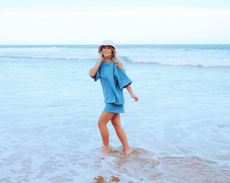 May include: A woman wearing a light blue, short-sleeved, oversized shirt and matching shorts walks on a sandy beach with ocean waves in the background. She is wearing a white bucket hat and smiling.