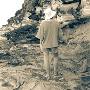 May include: A person wearing a white hat and a light brown shirt stands on a rocky beach. The person is looking out at the ocean.