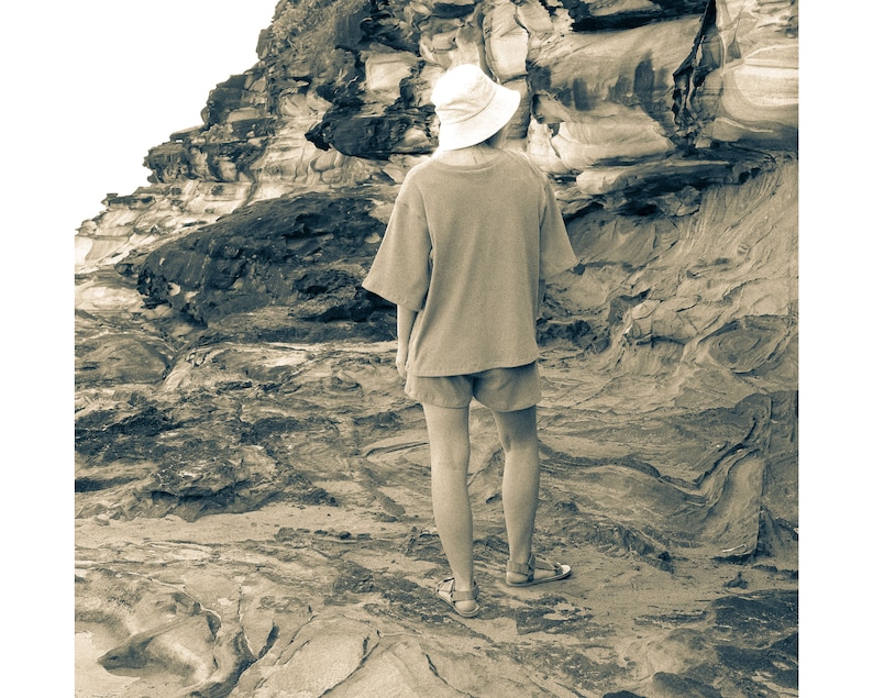 May include: A person wearing a white hat and a light brown shirt stands on a rocky beach. The person is looking out at the ocean.