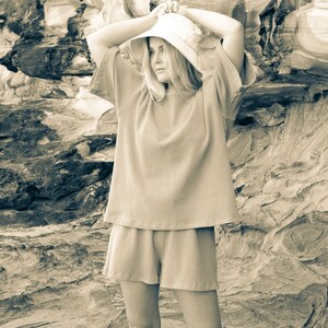 May include: A woman wearing a light brown, loose-fitting, short-sleeved top and matching shorts. She is wearing a light brown bucket hat and is standing in front of a rocky background.