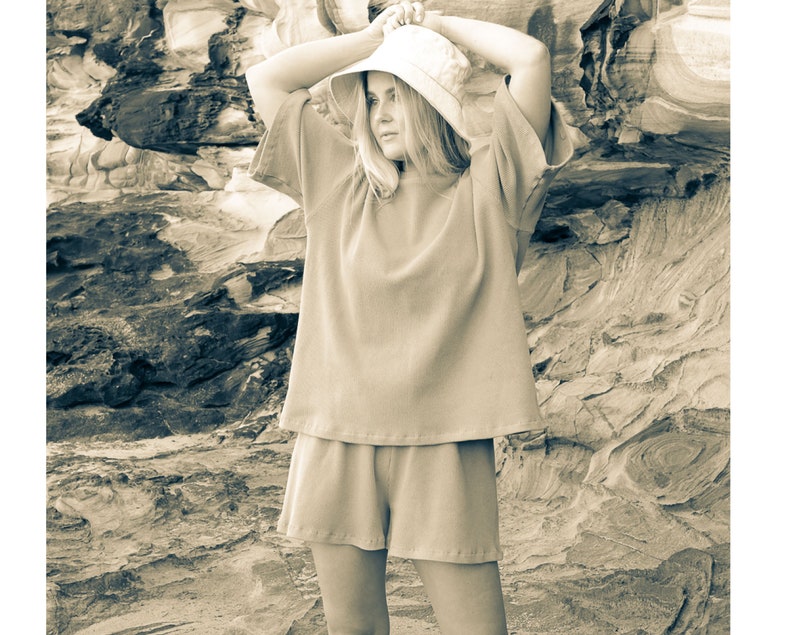 May include: A woman wearing a light brown, loose-fitting, short-sleeved top and matching shorts. She is wearing a light brown bucket hat and is standing in front of a rocky background.
