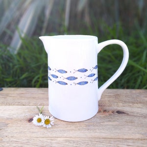 Può includere: Brocca in ceramica bianca con un design di pesci blu e marroni. La brocca ha un'ampia apertura e un manico.