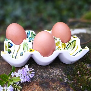 May include: A white ceramic egg holder with a green and yellow floral design. Three brown eggs are sitting in the holder.