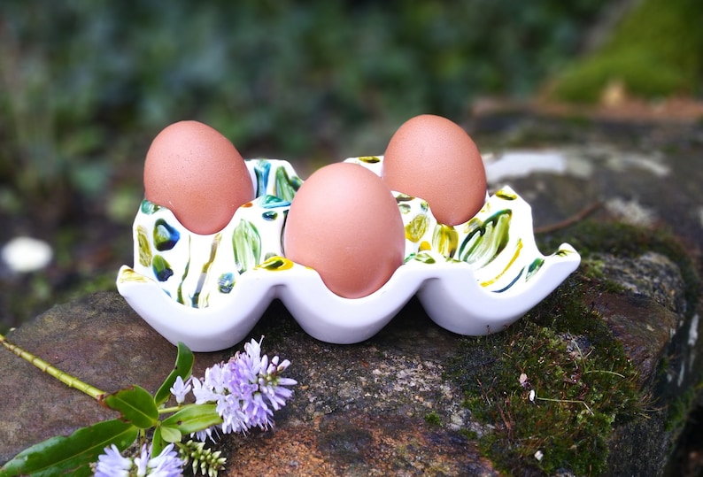 May include: A white ceramic egg holder with a green and yellow floral design. Three brown eggs are sitting in the holder.