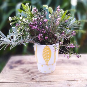 Peut inclure: Un vase en céramique blanche avec un motif géométrique jaune et blanc contient un bouquet de fleurs roses et blanches. Le vase a un fond arrondi et une large ouverture.