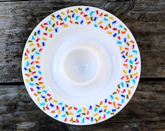 Puede incluir: Un huevero de cerámica blanca con un diseño de peces coloridos alrededor del borde. Los peces son en tonos de azul, verde, amarillo, naranja y rojo.