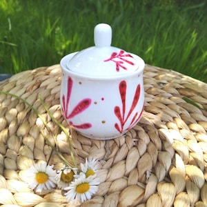 Peut inclure: Un bol à sucre en céramique blanche avec un couvercle, peint à la main avec des motifs floraux roses et dorés. Le bol est posé sur une surface en osier tressé.