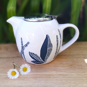 Peut inclure: Théière en céramique blanche avec un motif de feuilles bleu et blanc. La théière a un couvercle en acier inoxydable et un bec verseur. Deux marguerites blanches sont au premier plan.