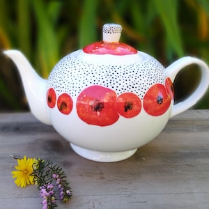Op de afbeelding: Witte theepot met een zwart-wit stippenpatroon en een rood klaproosontwerp.