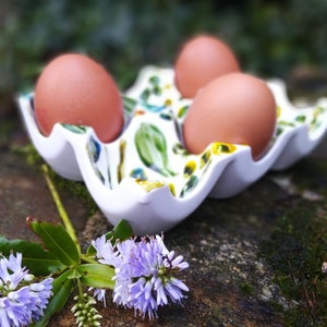 May include: Three brown eggs in a white ceramic egg holder with a floral design. The holder is sitting on a rock covered in moss.
