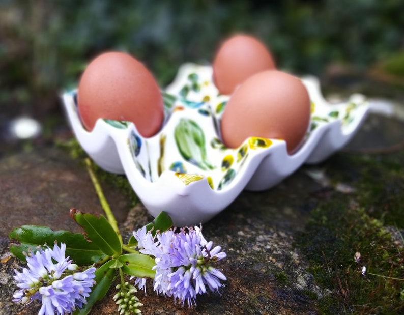 May include: Three brown eggs in a white ceramic egg holder with a floral design. The holder is sitting on a rock covered in moss.