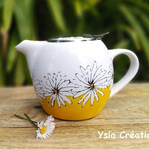 Puede incluir: Tetera de cerámica blanca con un diseño de margaritas amarillas y blancas. La tetera tiene una tapa de acero inoxidable con una pequeña asa.